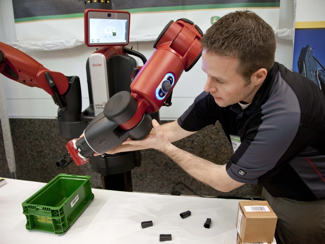 Worker demonstrating a parts-handling task beside a Baxter robot in a manufacturing setting